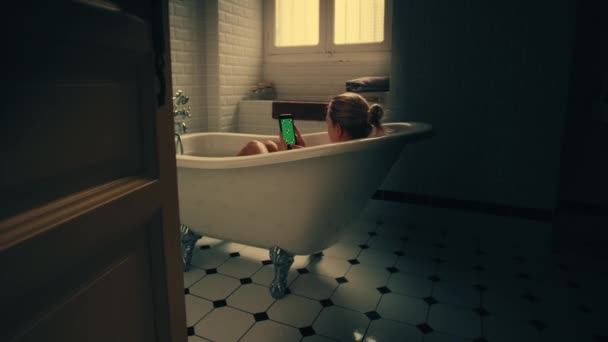 美しいブロンドの女の子は、リラックスしながら、緑のモックアップ画面のスマートフォンを使用してお風呂を取ります。魅力的な女の子は、ソーシャルメディアを閲覧し、ビデオを見てクロマキー携帯電話を使用しています。夕方のゴールデンアワーショット — ストック動画
