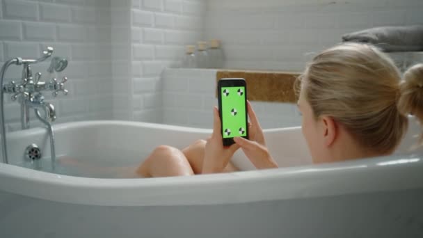 아름다운 금발소녀는 목욕을 하는 동안 녹색 모형 화면 스마트 폰을 사용합니다. 흑백 체커 타일이 있는 매력적인 빅토리아 스타일의 목욕 — 비디오