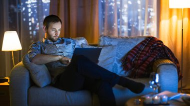 Akşam orta yaşlı adam oturur tutan bir kanepe laptop üstünde onun diz ve It.Tv eserlerinden mevcuttur üzerinde ve bu aydınlatan Him.Skyscrapers penceresinden görülür.