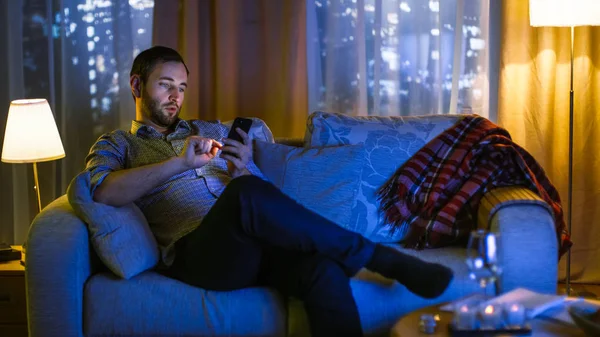 In the Evening Middle Aged Man Sits on a Sofa Uses Smartphone. Encendió la televisión lo ilumina. Vista de fondo de una gran ciudad con rascacielos . — Foto de Stock