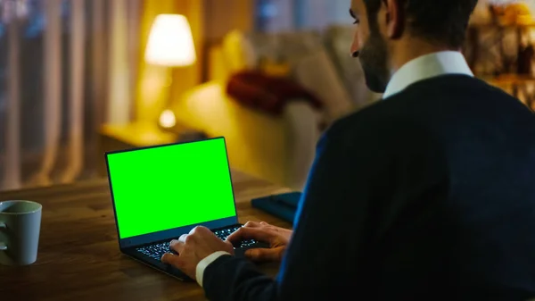 Daheim die Rückansicht eines Mannes, der an seinem Schreibtisch sitzt und auf einem Laptop mit grünem Bildschirm tippt. seine Wohnung ist in gelben Farben gehalten und warm. — Stockfoto
