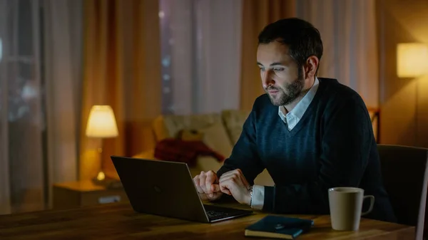 Respectable regardant l'homme du Moyen Âge travaille sur un ordinateur portable de sa maison. Son appartement a vue sur une grande ville. À l'intérieur, c'est chaleureux et confortable . — Photo