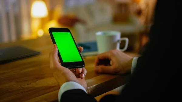 Otthon ember ül a asztal és használ Smartphone-val zöld képernyő rajta. Iszik pohárból. A lakásban történik, sárga színben és meleg. — Stock Fotó