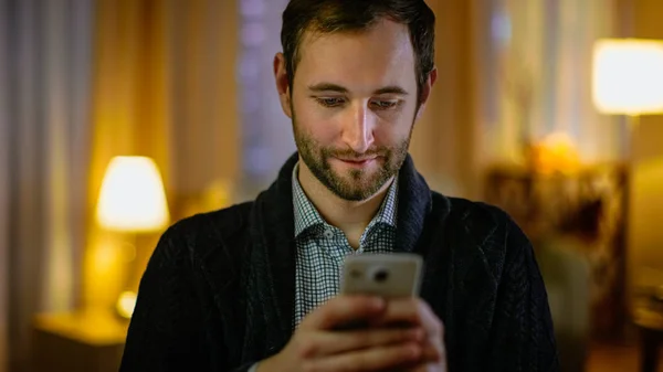 Porträt eines Mannes zu Hause, der sein Smartphone mit beiden Händen hält. Seine Wohnung ist in warmen Gelbtönen gehalten. Licht an. — Stockfoto