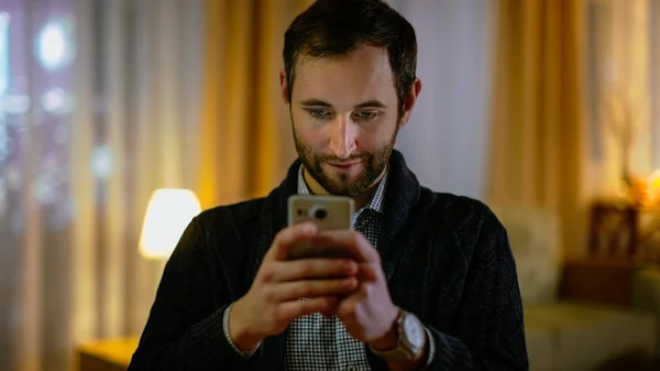 Porträt eines Mannes zu Hause, der sein Smartphone mit beiden Händen hält. Seine Wohnung ist in warmen Gelbtönen gehalten. Licht an. — Stockfoto
