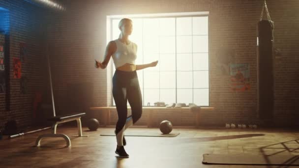 Fit αθλητική ξανθιά γυναίκα ασκήσεις με άλμα σχοινί σε ένα βιομηχανικό γυμναστήριο στιλ σοφίτας. Είναι συγκεντρωμένη στο πρόγραμμα εντατικής γυμναστικής της. Η εγκατάσταση έχει αφίσες κινήτρων στον τοίχο. — Αρχείο Βίντεο