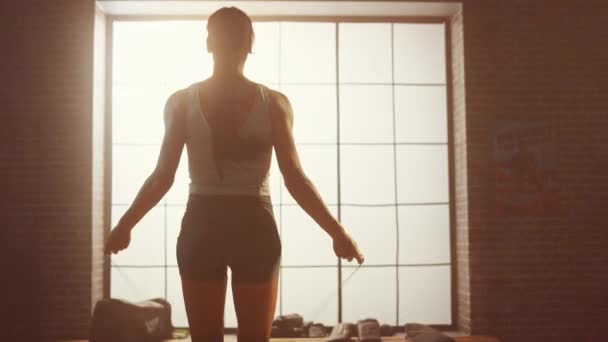 Sterke atletische vrouw oefeningen met Jumping touw in een loft-stijl industriële Gym. Ze concentreert zich op haar intense cross fitness trainingsprogramma. Gericht op groot raam. Back shot. — Stockvideo