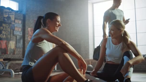 Zwei schön durchtrainierte, athletische Mädchen sitzen auf dem Boden einer Industrielokal-Turnhalle. Sie sind mit ihrem Trainingsprogramm zufrieden und geben erfolgreich eine hohe Fünf. starker männlicher Mann tritt in den Hintergrund. — Stockvideo