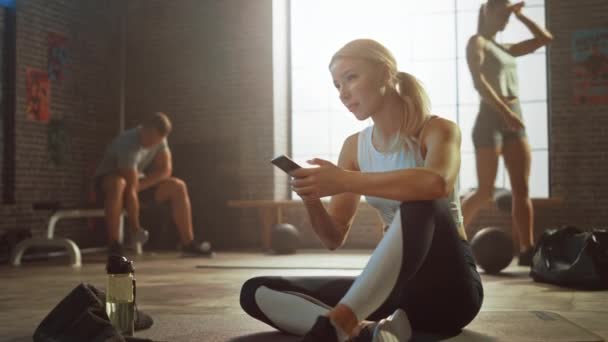 Glückliche und lächelnde schöne athletische junge Frau benutzt ein Smartphone, während sie auf einem Boden in einem Loft-Fitnessstudio sitzt. Sie tippt eine Nachricht und denkt nach. Ein Mann übt im Hintergrund. — Stockvideo