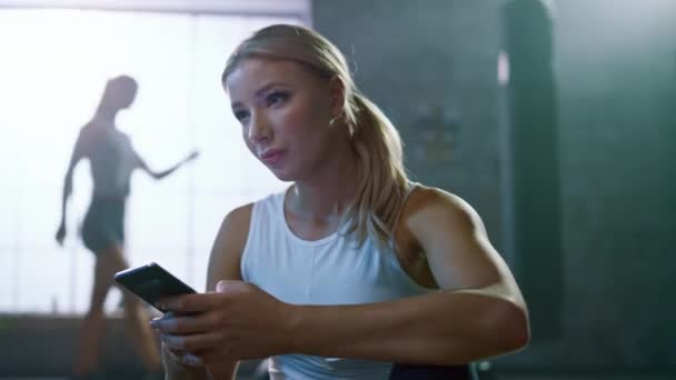 Selbstbewusste, schöne, athletische junge Frau benutzt ein Smartphone, während sie auf dem Boden in einem Loft-Fitnessstudio sitzt. Sie tippt eine Nachricht und denkt nach. Ein Mann übt im Hintergrund. — Stockvideo