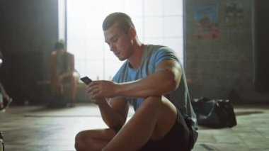 Yakışıklı ve Erkeksi Atletik Genç Adam Bir Loft Spor Salonu'nda Bir Katta Otururken Bir Akıllı Telefon kullanıyor. Mesaj yazıp düşünüyor. Alnında Ter var. Arka Planda Kadın Egzersiz.