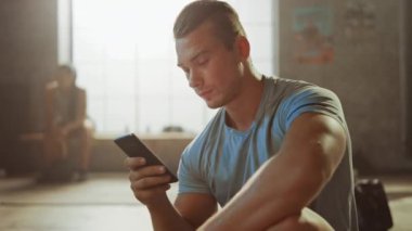 Yakışıklı ve Erkeksi Atletik Genç Adam Bir Loft Spor Salonu'nda Bir Katta Otururken Bir Akıllı Telefon kullanıyor. Mesaj yazıp düşünüyor. Alnında Ter var. Arka Planda Kadın Egzersiz.