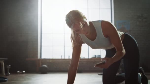 Krásná a mladá dívka používá smartphone aplikaci k instalačnímu programu pro její cvičení a začíná běžet na její fitness rohoži. Sportovní žena dělá horolezectví ve stylovém hardcore tělocvičně — Stock video