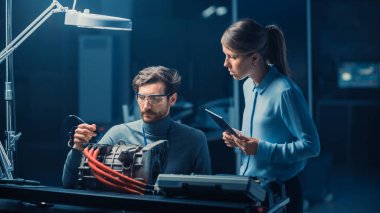 Tablet Bilgisayar ve Muayene Araçları ile Erkek ve Kadın Otomotiv Mühendisleri Bir Konsept Otomobil Şasi ile Yüksek Teknoloji Laboratuvarında Bir Elektrik Motoru Test Ederken Bir Konuşma Yapıyor.
