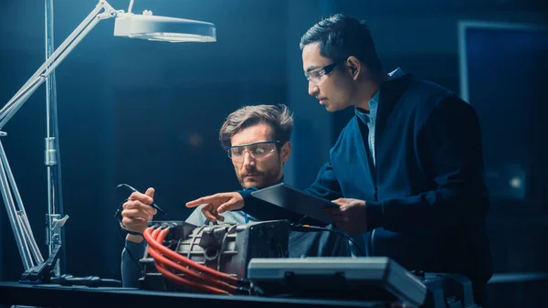 Two Professional Automotive Engineers with a Tablet Computer and Inspection Tools are Having a Conversation While Testing an Electric Engine in a High Tech Laboratory with a Concept Car Chassis.