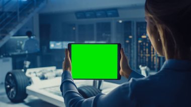 Kadın Mühendis Yeşil Ekran Mock Up ile Tablet Bilgisayar Tutar ve Yüksek Teknoloji Geliştirme Laboratuvarı'nda Tekerlekler, Piller ve Motor Ayakta Elektrikli Araba Şasi Prototip bakar.