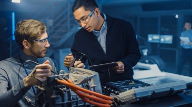Tablet Bilgisayar ve Denetim Araçları ile İki Profesyonel Otomotiv Mühendisleri Bir Konsept Otomobil Şasi ile Yüksek Teknoloji Laboratuvarında Bir Elektrik Motoru Test Ederken Bir Konuşma Yapıyor.