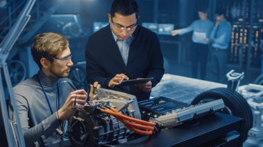 Tablet Bilgisayar ve Denetim Araçları ile İki Profesyonel Otomotiv Mühendisleri Bir Konsept Otomobil Şasi ile Yüksek Teknoloji Laboratuvarında Bir Elektrik Motoru Test Ederken Bir Konuşma Yapıyor.