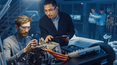 Tablet Bilgisayar ve Denetim Araçları ile İki Profesyonel Otomotiv Mühendisleri Bir Konsept Otomobil Şasi ile Yüksek Teknoloji Laboratuvarında Bir Elektrik Motoru Test Ederken Bir Konuşma Yapıyor.