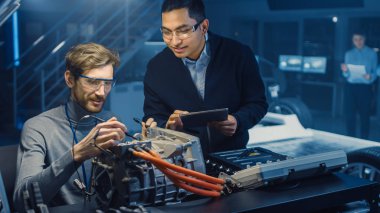 Tablet Bilgisayar ve Denetim Araçları ile İki Profesyonel Otomotiv Mühendisleri Bir Konsept Otomobil Şasi ile Yüksek Teknoloji Laboratuvarında Bir Elektrik Motoru Test Ederken Bir Konuşma Yapıyor.