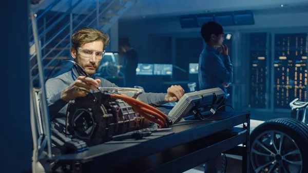 Ingénieur automobile professionnel en lunettes avec un ordinateur et des outils d'inspection teste un moteur électrique usagé dans un laboratoire de haute technologie avec un châssis Concept Car . — Photo