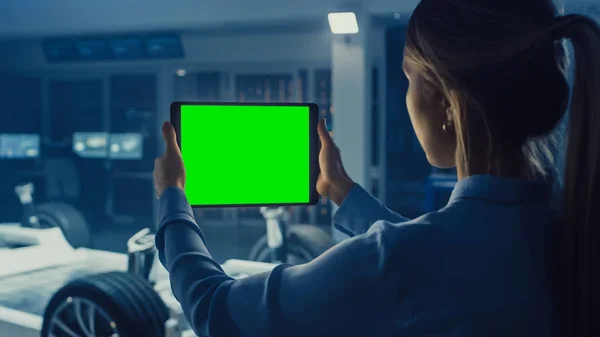 Engenheira Feminina detém um computador tablet com tela verde Mock Up e olha para o protótipo de chassi de carro elétrico com rodas, baterias e motor em pé em um laboratório de desenvolvimento de alta tecnologia . — Fotografia de Stock