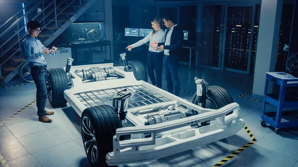 Vue à grand angle d'une salle de laboratoire de développement avec une équipe professionnelle diversifiée d'ingénieurs et de scientifiques travaillant sur un châssis de voiture électrique avec roues, batteries, moteur et suspension . — Photo