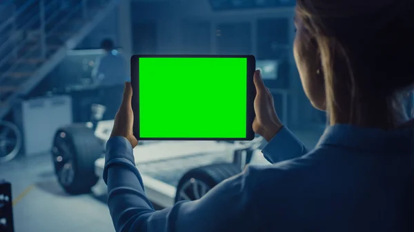Une ingénieure tient un ordinateur tablette avec écran vert maquillé et regarde le prototype de châssis de voiture électrique avec roues, batteries et moteur debout dans un laboratoire de développement de haute technologie . — Photo