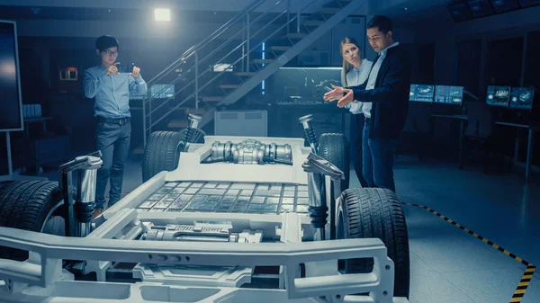 Automotive ontwerpingenieurs praten tijdens het werken aan elektrische auto chassis prototype. In innovatie laboratorium faciliteit concept voertuig frame omvat wielen, ophanging, motor en batterij. — Stockfoto