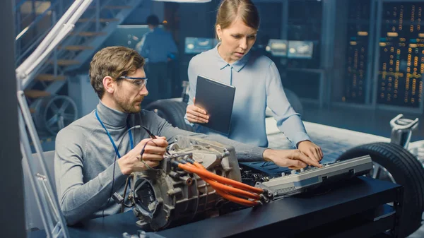Manliga och kvinnliga fordons ingenjörer med en Tablet PC och inspektionsverktyg har en konversation samtidigt testa en elektrisk motor i ett högteknologiskt laboratorium med ett konceptbil chassi. — Stockfoto