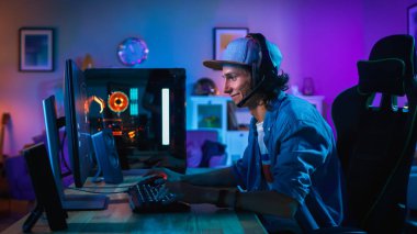 Gamer Mikrofon Ile Kulaklığı Koyar ve Kişisel Bilgisayarında Shooter Online Video Oyunu Oynamaya Başlar. Oda ve Pc Renkli Neon Led Işıklar var. Genç Adam Şapka Takıyor. Evde Rahat Akşam.