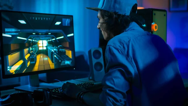 Emocionado jugador jugando en primera persona Shooter juego de video en línea en su potente ordenador personal. La habitación y la PC tienen luces led de neón coloridas. El joven lleva una gorra. Acogedora noche en casa . —  Fotos de Stock