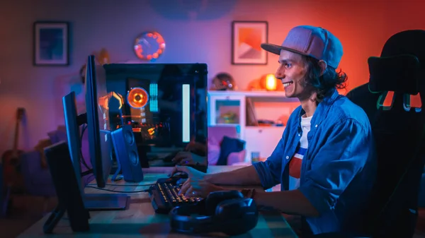 Jogador animado jogando jogo de tiro em primeira pessoa jogo de vídeo on-line em seu computador pessoal. Quarto e PC têm luzes LED de néon coloridas. O Jovem está a usar um boné. Noite aconchegante em casa . — Fotografia de Stock
