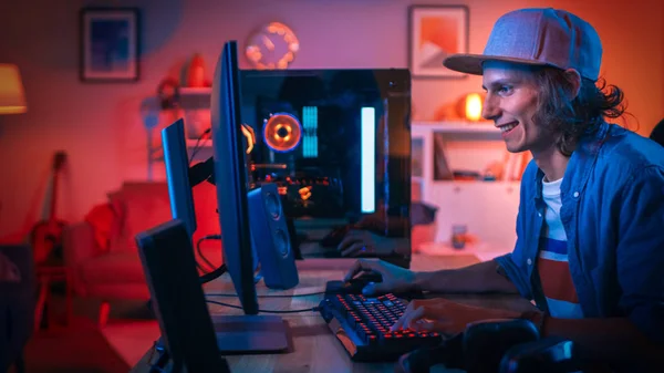 Jugador profesional que juega el videojuego en línea del tirador en primera persona en su computadora personal potente con luces led de neón coloridas. El joven lleva una gorra. Salón con Lámparas Naranjas Cálidas. Buenas noches. . — Foto de Stock