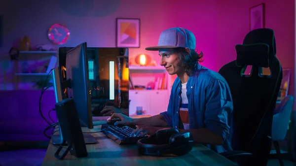 Jugador profesional que juega el videojuego en línea del tirador en primera persona en su computadora personal potente con luces led de neón coloridas. El joven lleva una gorra. Salón Lit con Lámparas Rosa. Buenas noches. . — Foto de Stock