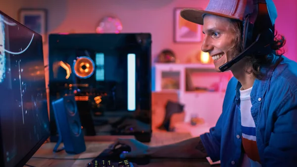 Gamer pone sus auriculares con un micrófono y comienza a jugar Shooter Juego de vídeo en línea en su ordenador personal. La habitación y la PC tienen luces led de neón coloridas. El joven lleva una gorra. Acogedora noche en casa . — Foto de Stock