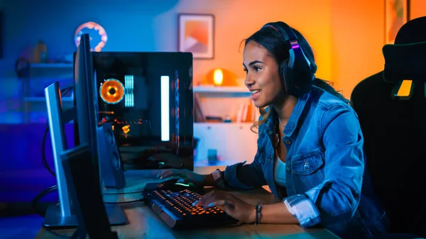 Hübsches und aufgeregtes schwarzes Spielermädchen mit Kopfhörern spielt Ego-Shooter-Online-Videospiel auf ihrem Computer. Zimmer und PC haben bunte Neon-LED-Lichter. Gemütlicher Abend zu Hause. — Stockfoto