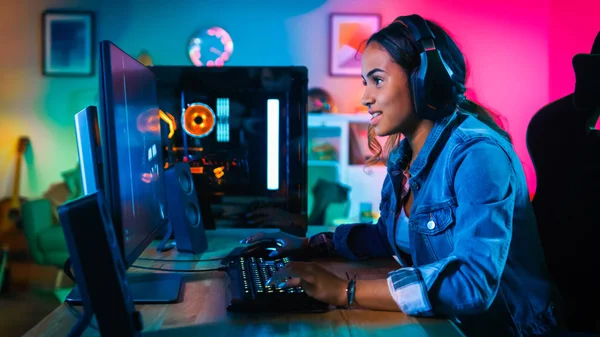 Retrato de una guapa y excitada jugadora negra con auriculares sonriendo a la cámara. Ella está sentada en la silla al lado de la computadora personal potente. Inicio y PC es Lit con luces de neón . —  Fotos de Stock