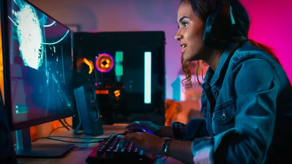 Krásná a vzrušená černá Gamer Girl ve sluchátkách hraje první osobu v online videohře na jejím počítači. Pokoje a počítač mají barevná neonová světla. Útulný večer doma. — Stock fotografie