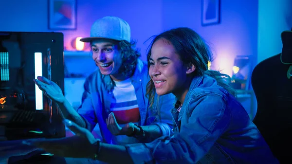 Förvånad och upphetsad ung man och bloggare flicka tittar på en åtgärd video på en kraftfull persondator. De är aktivt Gesturing. Rummet och datorn har färgglada Neonled-lampor. Mysig kväll hemma. — Stockfoto
