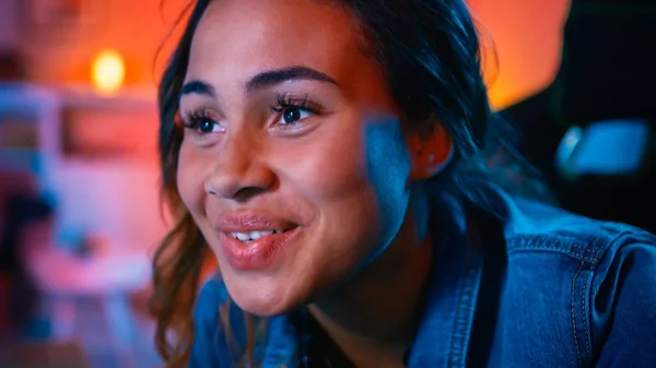 Close-up portret van een mooi opgewonden zwart meisje kijken naar een actie video op een computer. Ze heeft donker haar en bruine ogen. Scherm voegt reflecties toe aan haar gezicht. Gezellige kamer verlicht met warm rood licht. — Stockfoto