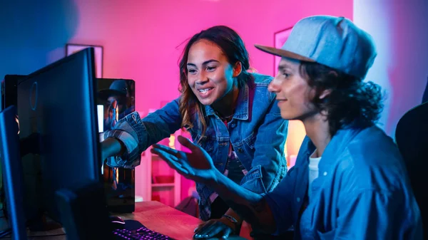 Twee blogger vrienden of een paar actief bespreken iets op een computer scherm. Mooi zwart meisje wijst naar het scherm en jonge man stemt in met haar creatieve ideeën. Gezellige kamer verlicht met ping neon licht. — Stockfoto