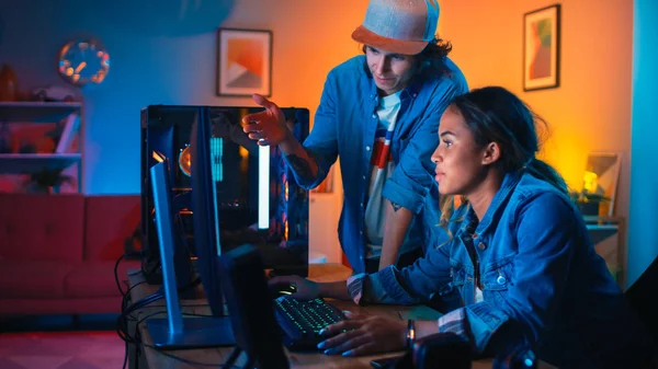 Dois amigos ou um casal discutem estratégias futuras em um jogo de computador. Pretty Black Girl mostra seu trabalho para um jovem e ele compartilha ideias criativas. Quarto acolhedor é lit com luz quente e néon . — Fotografia de Stock