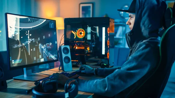 Professionell Gamer och streamer spelar första-person shooter online video spel på hans coola persondator. Den unge mannen bär mössa och huva. Rummet och datorn har färgglada Neonled-lampor. Mysig kväll hemma. — Stockfoto