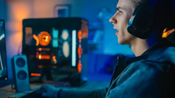 Szczęśliwy Gamer w zestawie słuchawkowym z mikrofonem gra strzelanka online wideo na jego komputerze osobistym. Daje rozkazy innym graczom. Pokój i komputer mają kolorowe Neon LED Lights. Przytulny wieczór w domu. — Zdjęcie stockowe