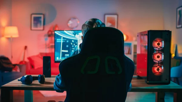 Tillbaka skott av en gamer spelar första person shooter online video spel på sin kraftfulla persondator. Rummet och datorn har varma färgglada Neonled-lampor. Mysig kväll hemma. — Stockfoto