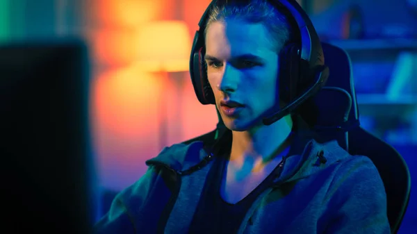 Gamer im Headset mit Mikrofon, das in einem Online-Videospiel auf seinem PC spielt. Er gibt anderen Spielern Kommandos. Zimmer und PC haben bunte Neon-LED-Lichter. Gemütlicher Abend zu Hause. — Stockfoto