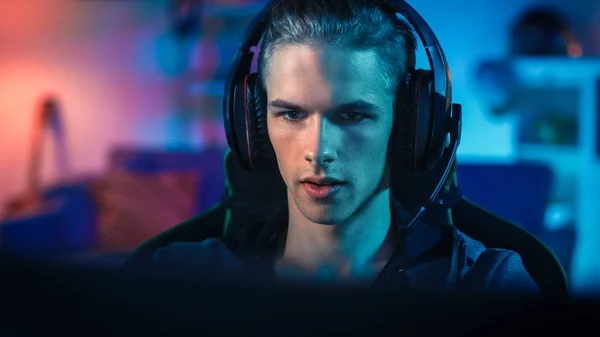 Gamer im Headset mit Mikrofon, der auf seinem PC ein Online-Videospiel spielt. Er konzentriert sich auf den Monitor. Zimmer und PC haben bunte Neonröhren. Gemütlicher Abend zu Hause. — Stockfoto