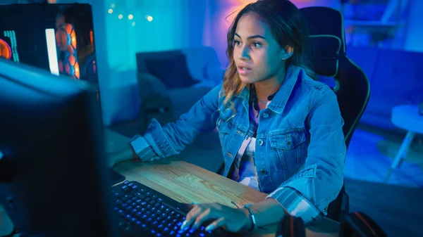 Aufgeregtes und hübsches schwarzes Spielermädchen spielt ein Online-Videospiel auf ihrem PC. Zimmer und PC haben bunte kalte Neonröhren. Gemütlicher Abend zu Hause. — Stockfoto