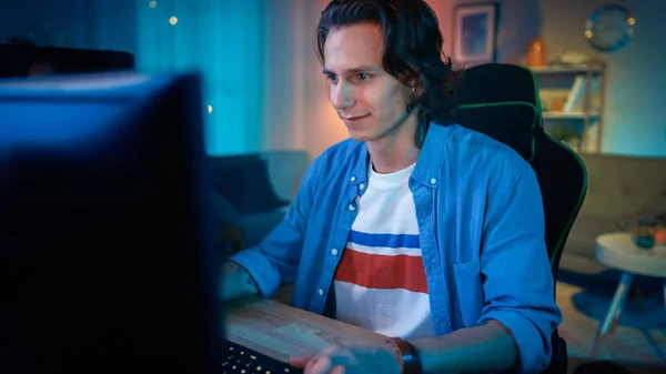 Emocionado jugador jugando videojuego en línea en su computadora personal. La habitación y la PC tienen luces led de neón cálidas coloridas. El joven tiene el pelo largo y la sonrisa guapa. Acogedora noche en casa . —  Fotos de Stock